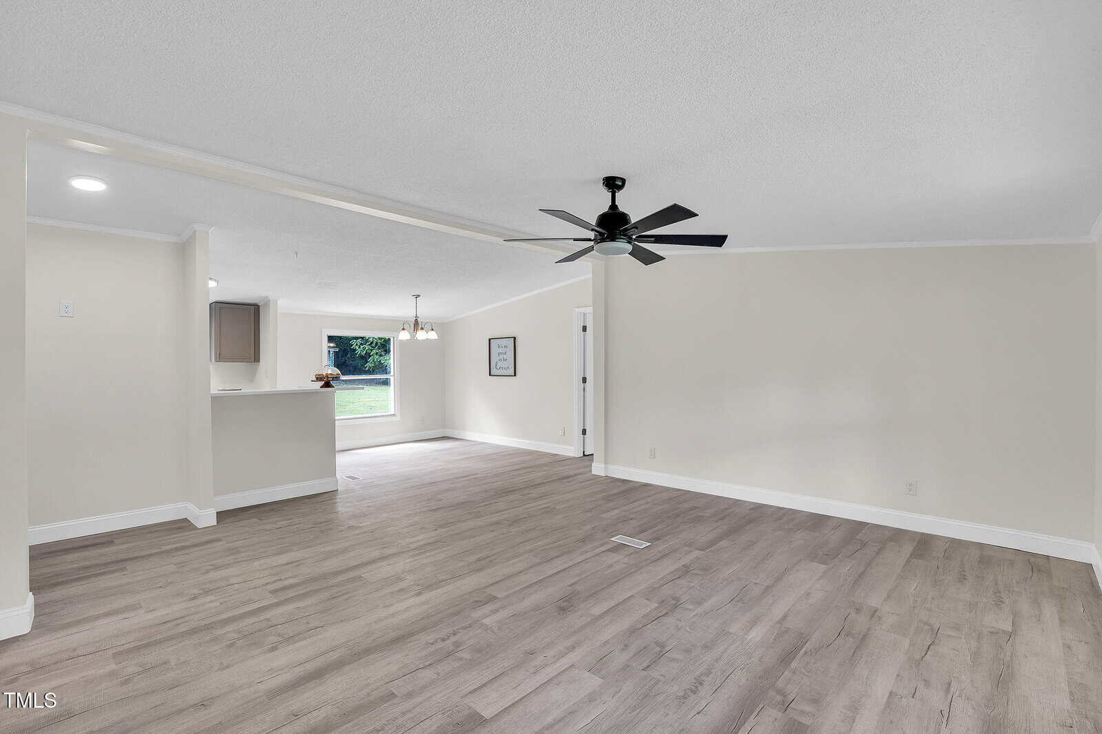 A spacious, empty room with light wood flooring, white walls, a black ceiling fan, and an open view into a kitchen area and dining nook with windows showing green grass outside.