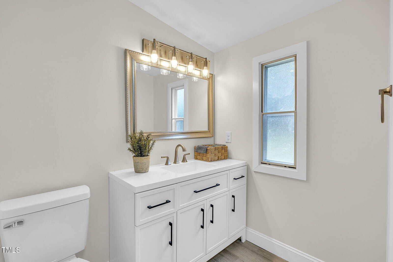 Modern bathroom with a white vanity, double sinks, brushed gold fixtures, a large framed mirror, five-light vanity fixture, frosted window, and a white toilet. A small potted plant decorates the countertop.