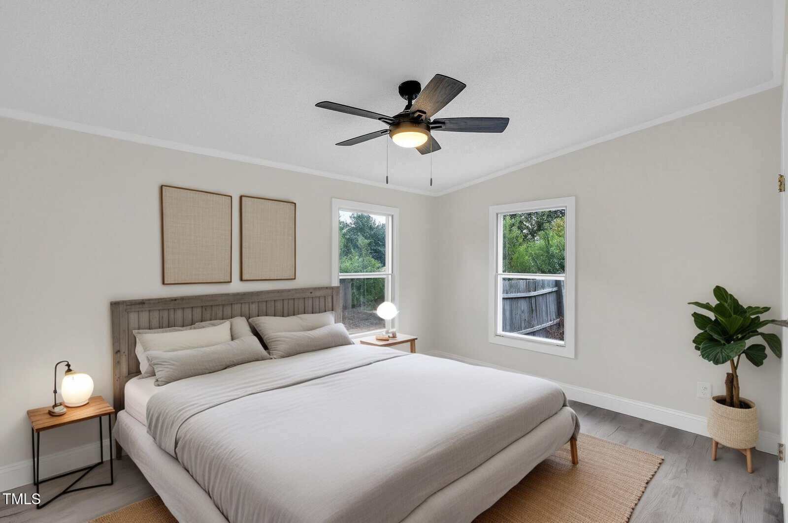 Minimalist bedroom with a large bed, neutral bedding, two wooden nightstands with lamps, wall art above the headboard, a ceiling fan, two windows, a green plant in the corner, and light-colored walls and flooring.