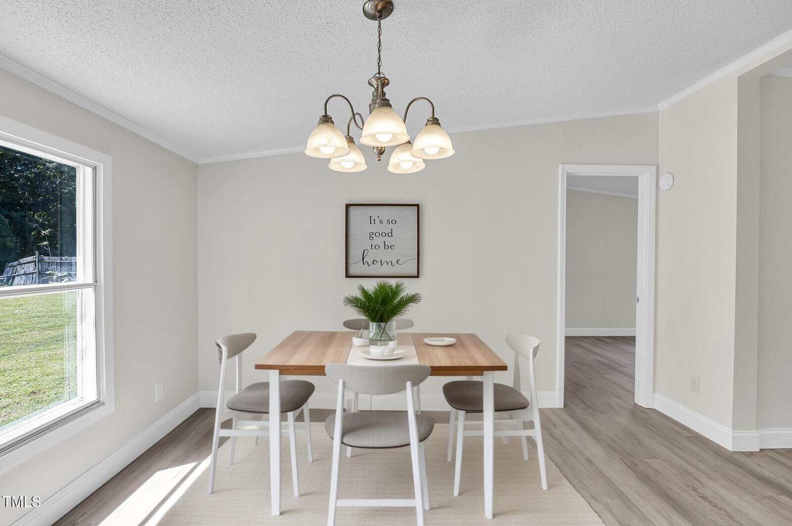 A dining room with a wooden table, four white chairs, a potted plant centerpiece, and a chandelier. A framed sign on the wall reads "It's so good to be home"; sunlight enters from a window on the left.