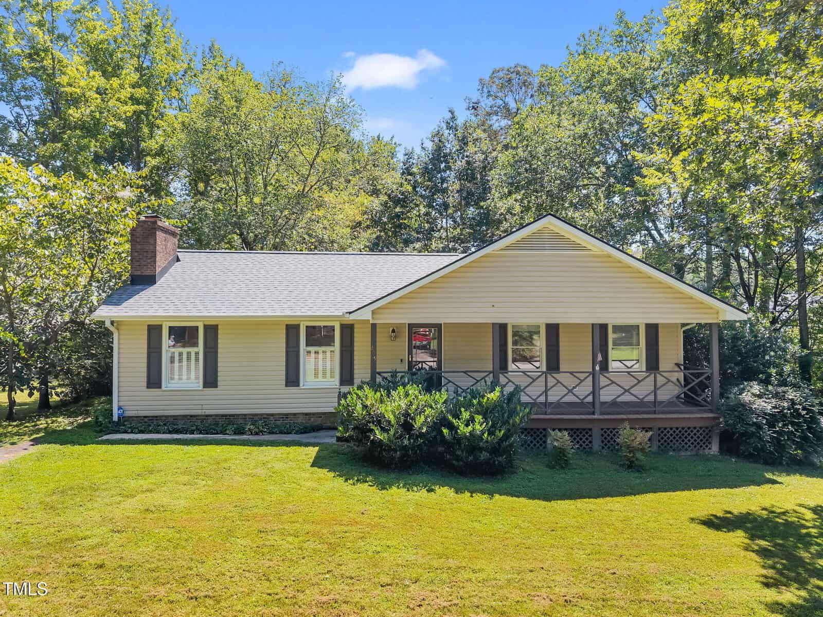 Charming single-story beige house with dark shutters and a covered front porch at 209 W Dogwood Drive in Creedmoor, set on a green lawn with bushes and surrounded by tall trees under a blue sky.