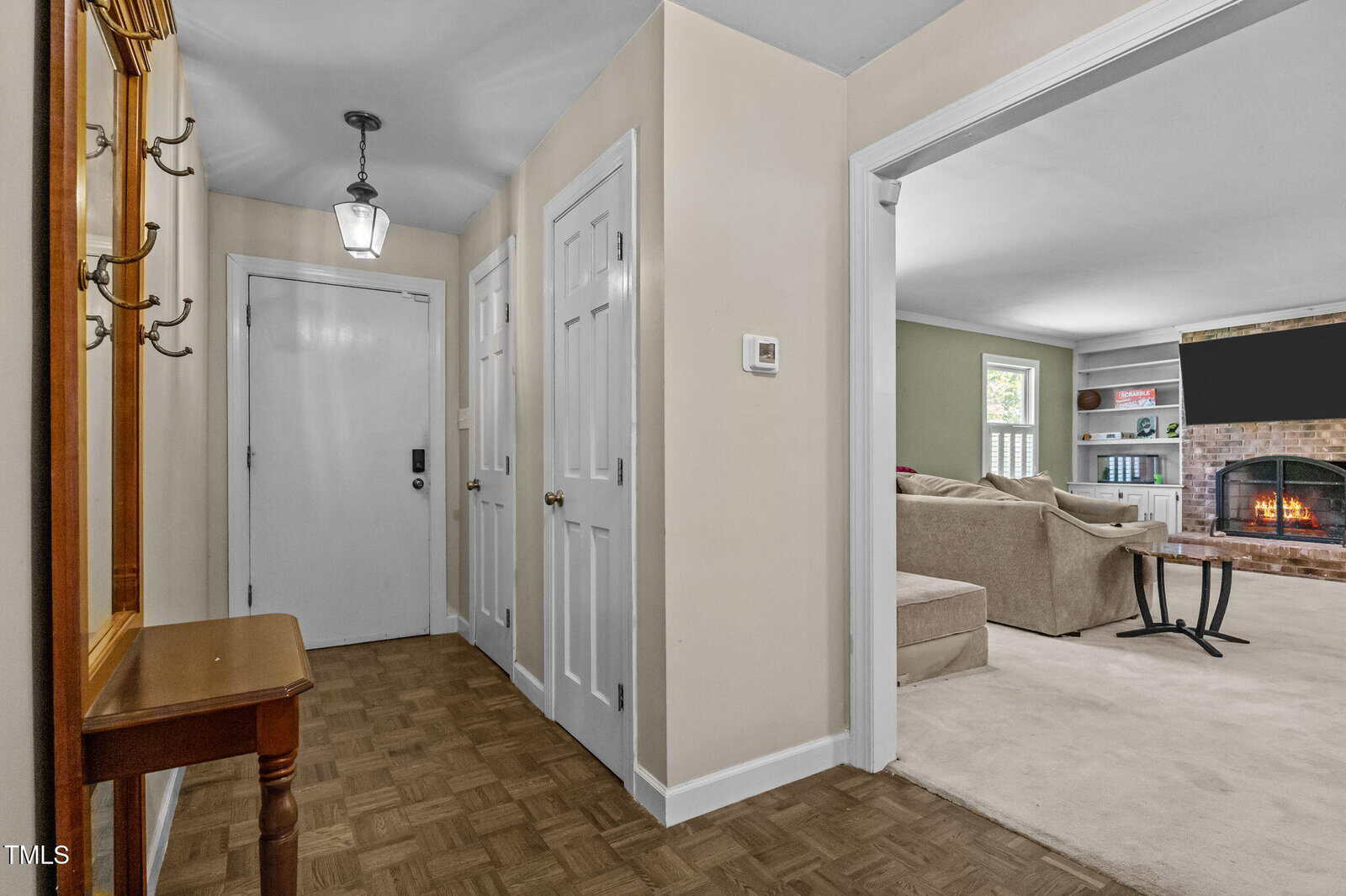 A hallway with beige walls and parquet flooring has a small wooden table and coat rack on the left. Three white doors are visible, and an open doorway leads to a living room with a fireplace and beige sectional sofa.