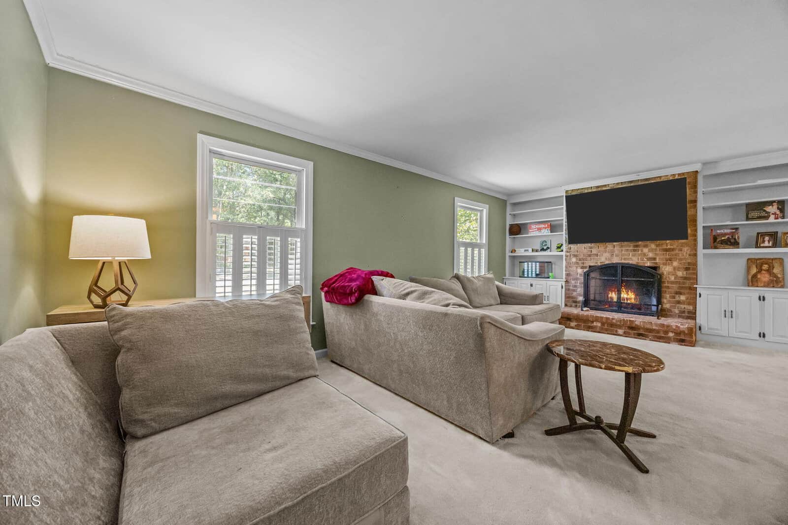 A living room with light-colored walls and carpet, beige sofas, a round wooden side table, built-in shelves, and a brick fireplace with a large TV above it. A table lamp is lit near two windows.