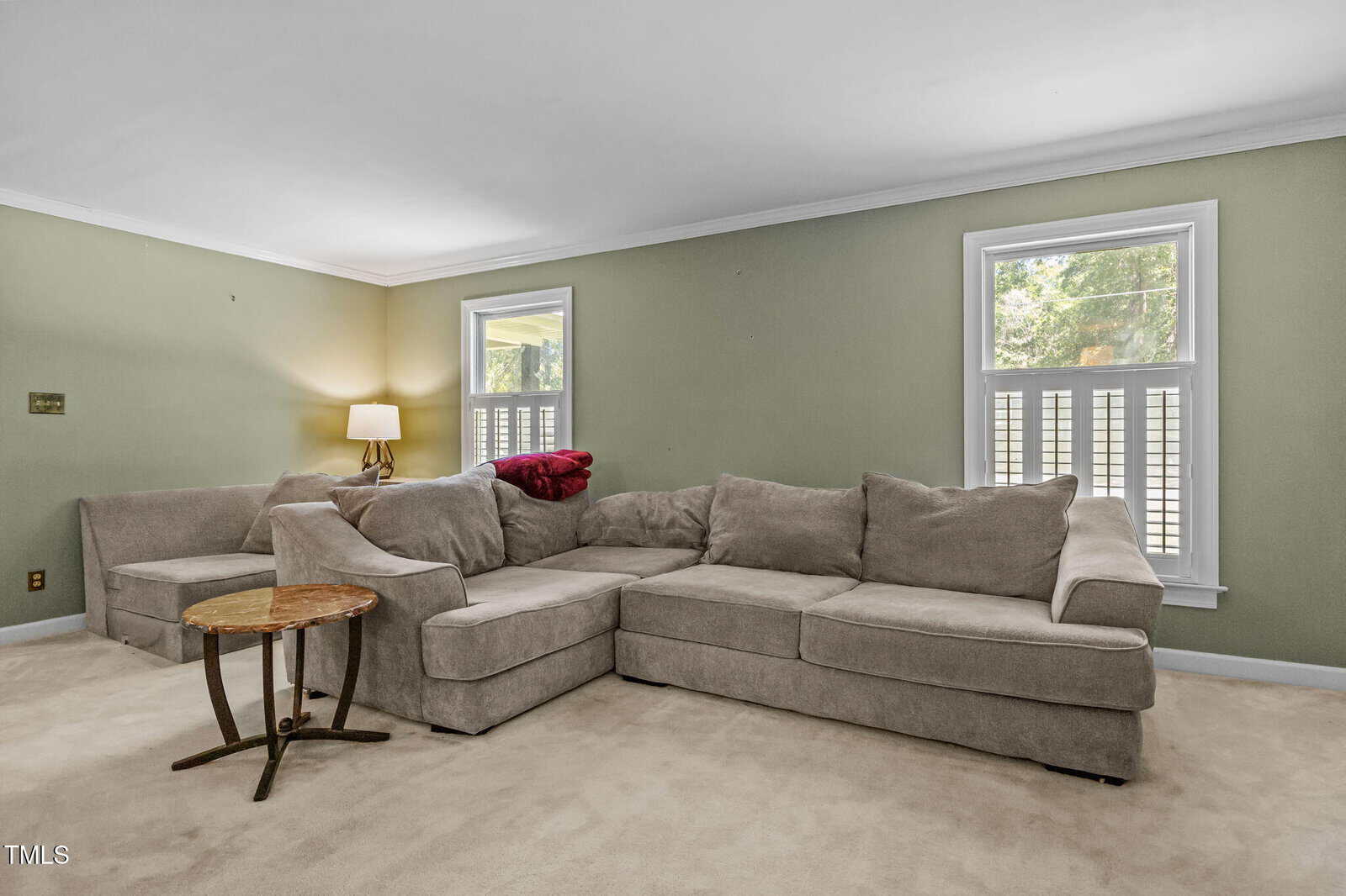 A living room with light green walls, beige carpet, a large beige sectional sofa, a small round wooden side table, a lamp, and two windows letting in natural light.