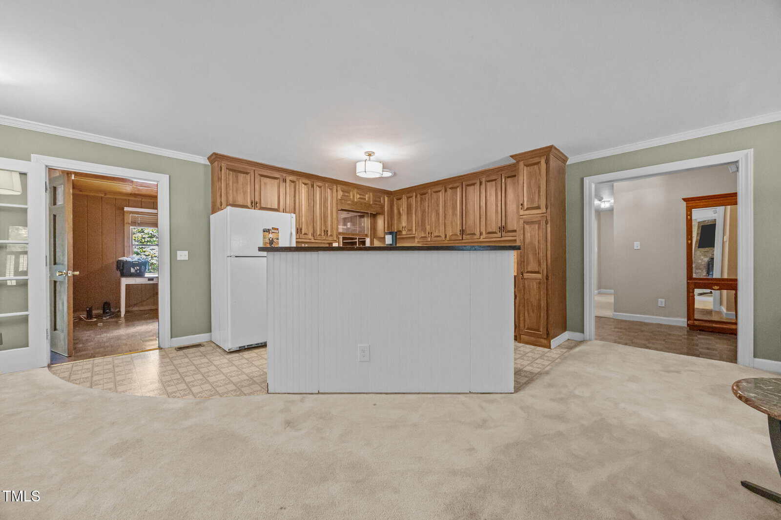 A kitchen with wood cabinets, white refrigerator, and raised counter is shown. It is bordered by two open doorways leading to other rooms, with a beige carpet and patterned linoleum flooring.