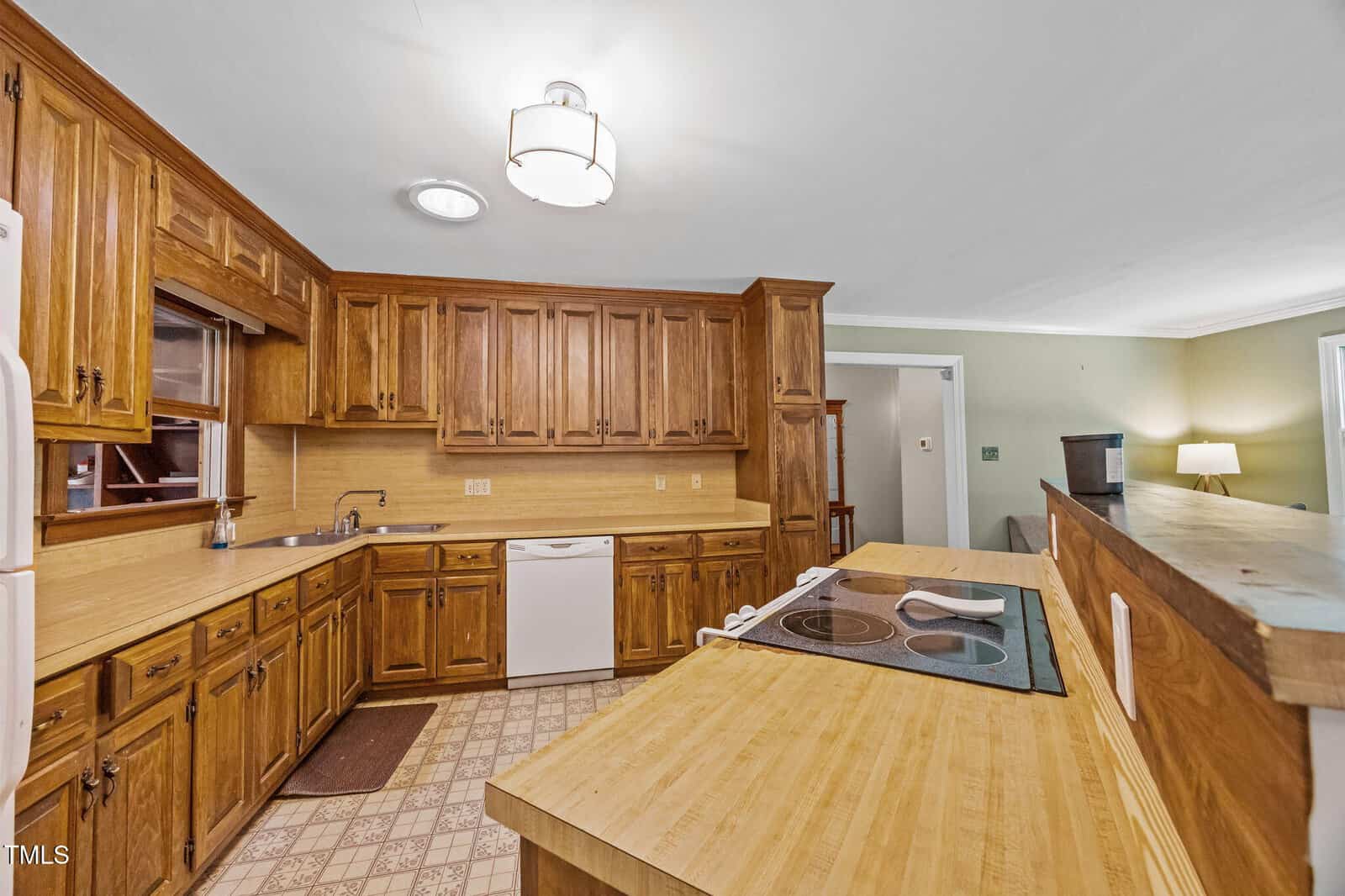 A kitchen with wooden cabinets, a white dishwasher, a white refrigerator, an electric cooktop on a wood counter island, and patterned tile flooring. The adjacent room is partially visible with green walls and a lamp.
