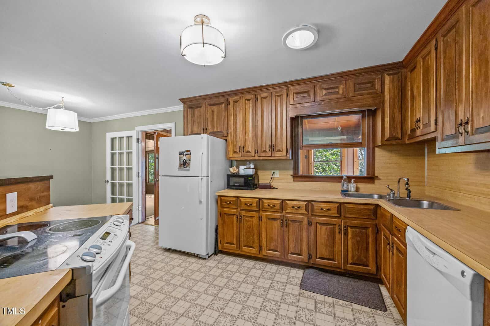 A kitchen with wooden cabinets, white appliances including a refrigerator, stove, and dishwasher, a double sink under a window, tile flooring, and ceiling light fixtures. A door with glass panels leads to another room.