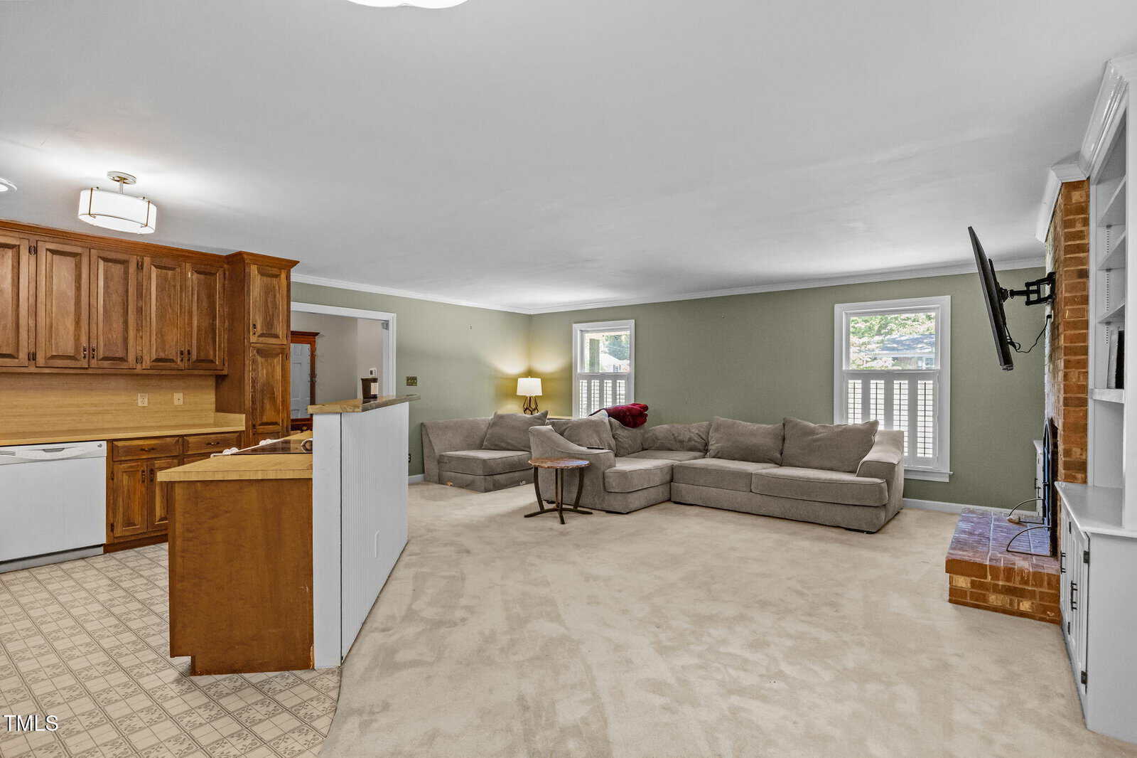 Open living area with beige carpet, light green walls, a large gray sectional sofa, wall-mounted TV above a brick hearth, and an adjacent kitchen with wood cabinets and white appliances. Multiple windows provide natural light.