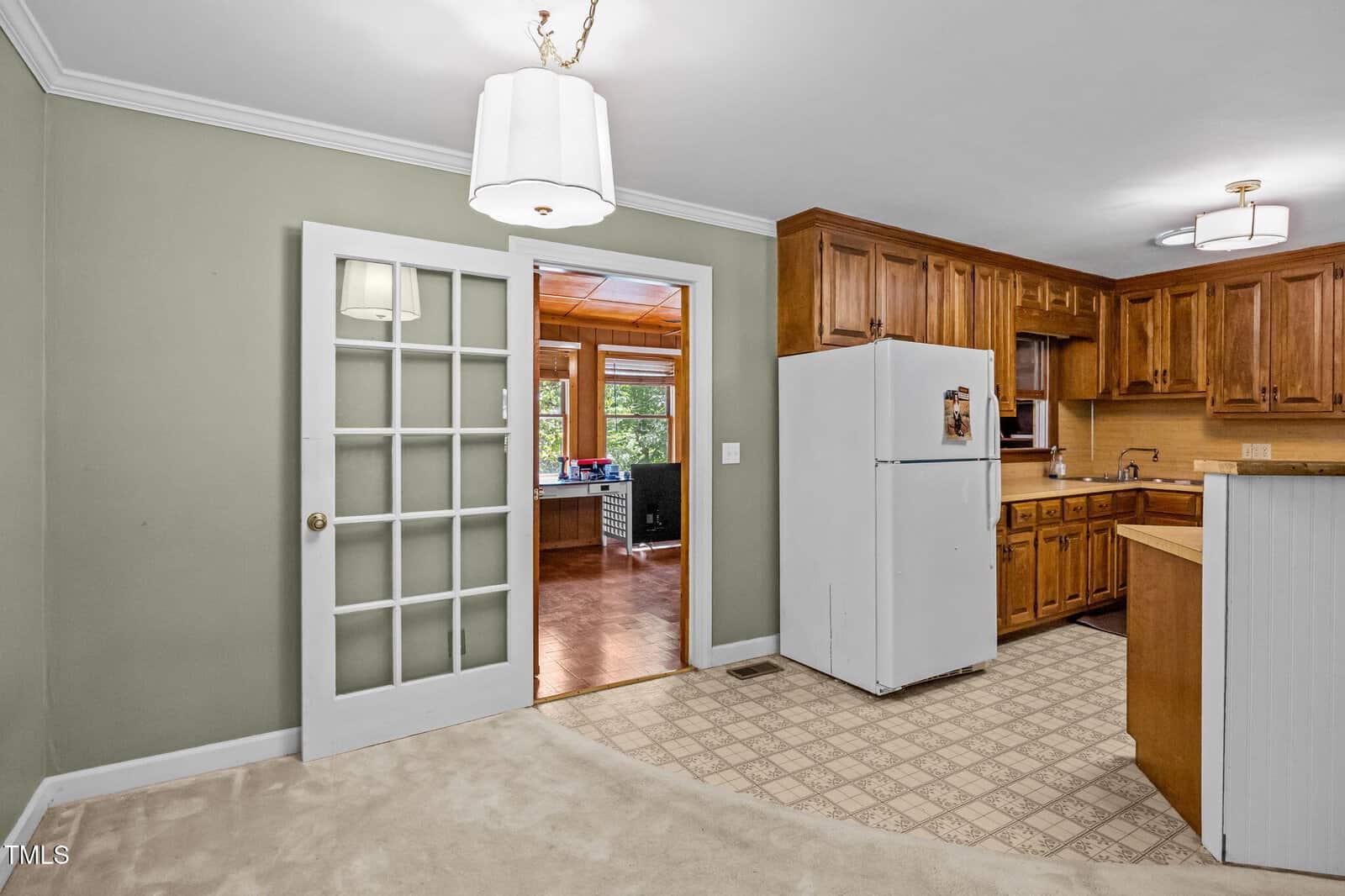A kitchen with light green walls, wood cabinets, a white refrigerator, and patterned linoleum flooring. A French door leads to another room with wood-paneled walls and large windows. Two ceiling light fixtures are visible.