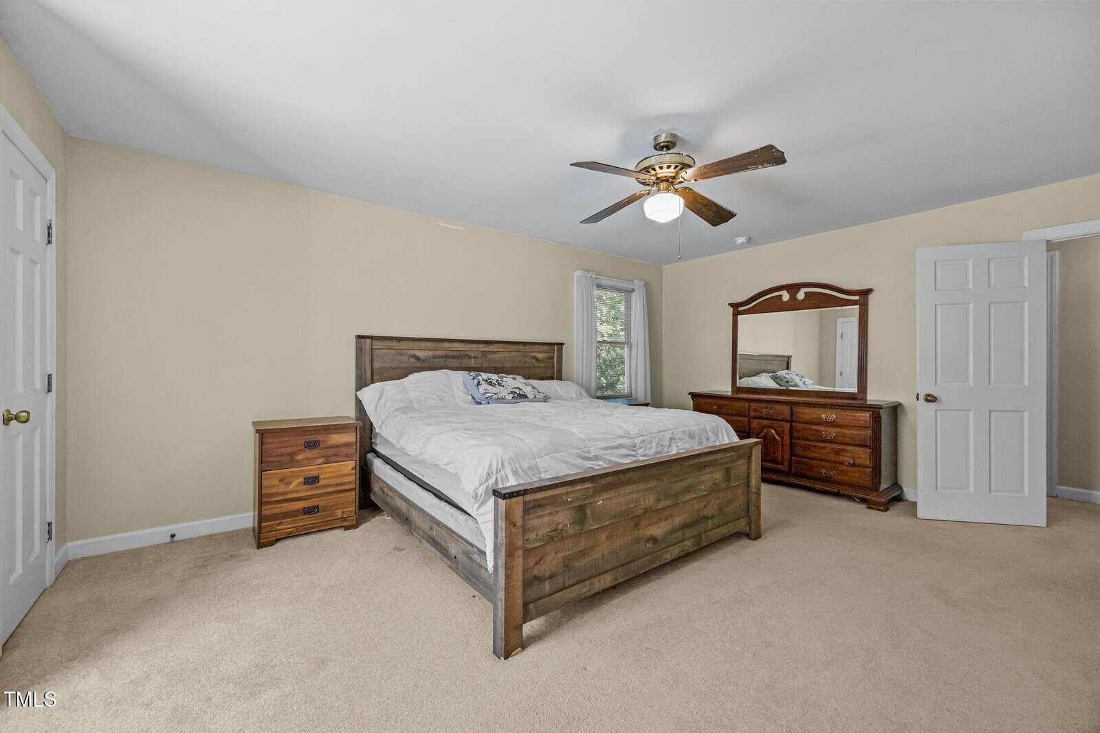 A bedroom with beige walls and carpet, featuring a wooden bed with white bedding, a matching wooden nightstand, a dresser with a mirror, two doors, a ceiling fan with a light, and a window.