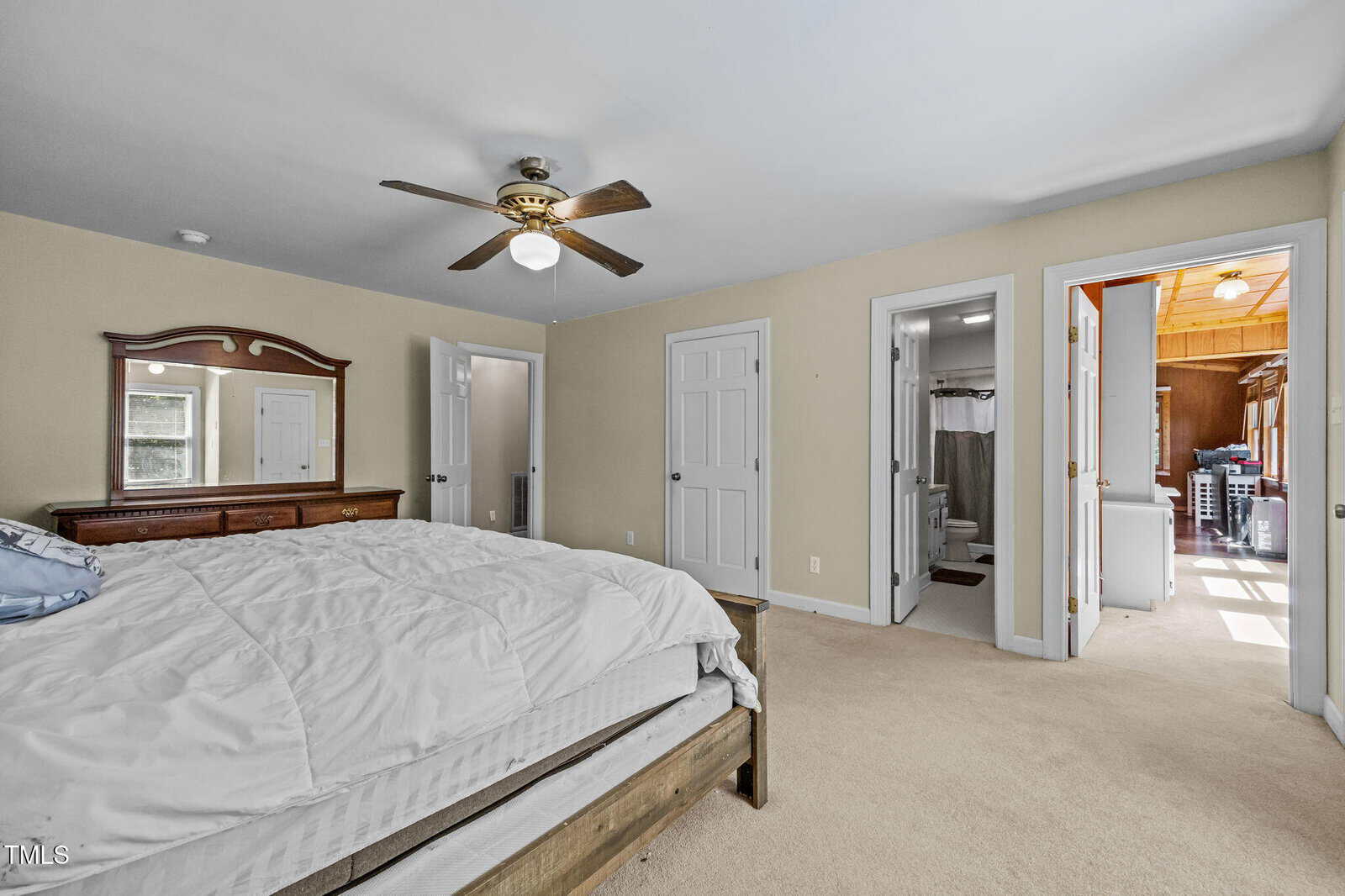 A spacious bedroom with a ceiling fan, large bed, dresser with mirror, beige walls, carpeted floor, and three doors leading to a bathroom, a closet, and another room. Natural light fills the space.