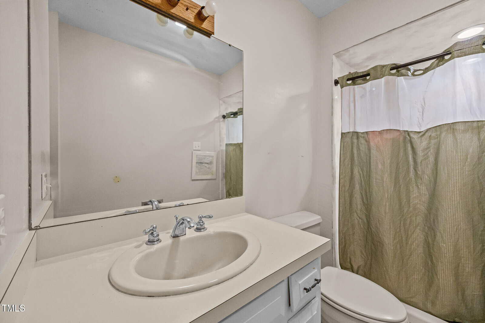 Small bathroom with a white sink and countertop, mirror above, toilet next to it, and a shower with a green and white curtain. Walls are light-colored, and lights are mounted above the mirror.