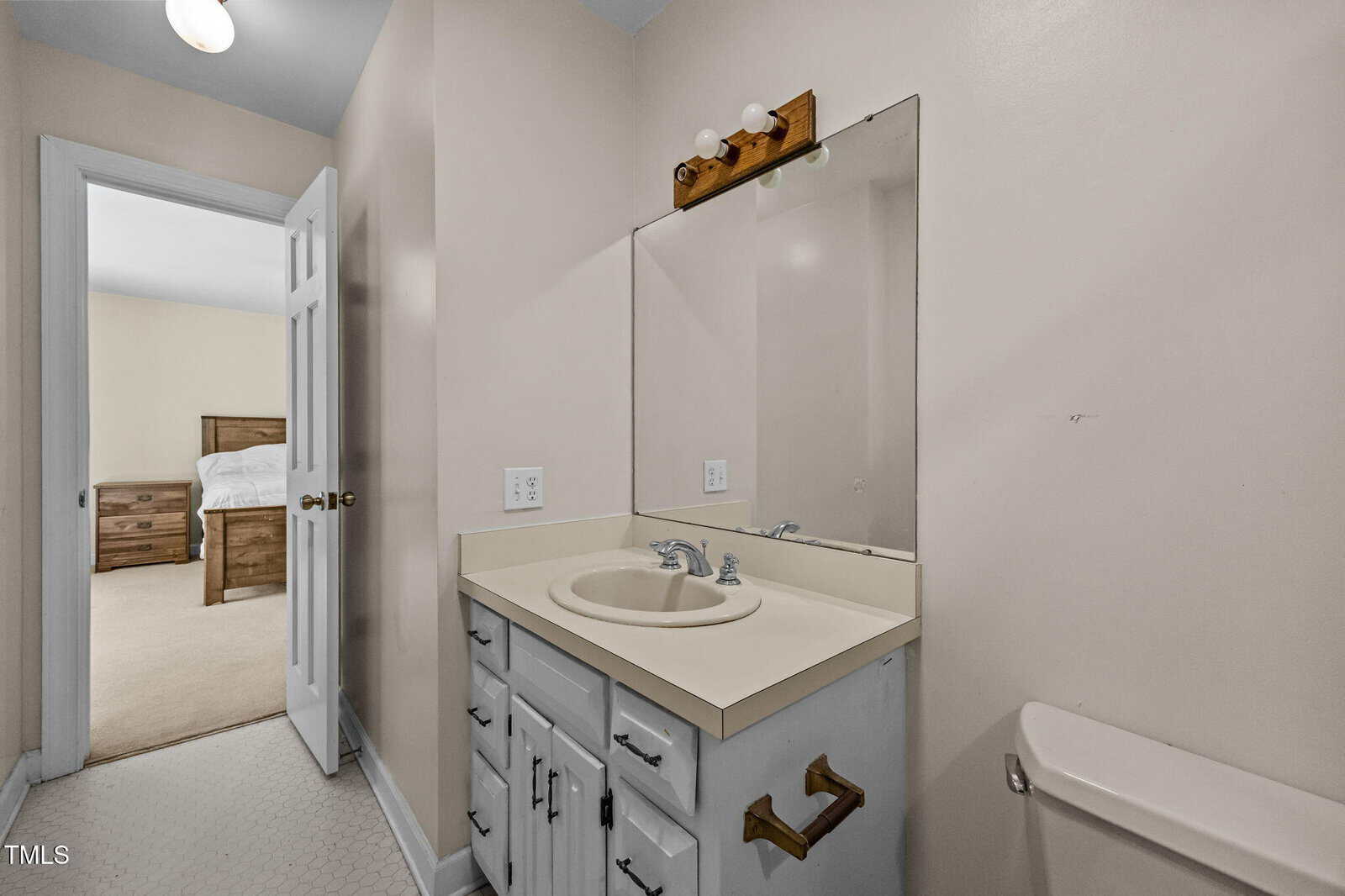 A bathroom with a single sink vanity, large mirror, and beige walls. A towel holder is mounted near the toilet. An open door leads to a bedroom with a wooden bed and beige carpet visible.