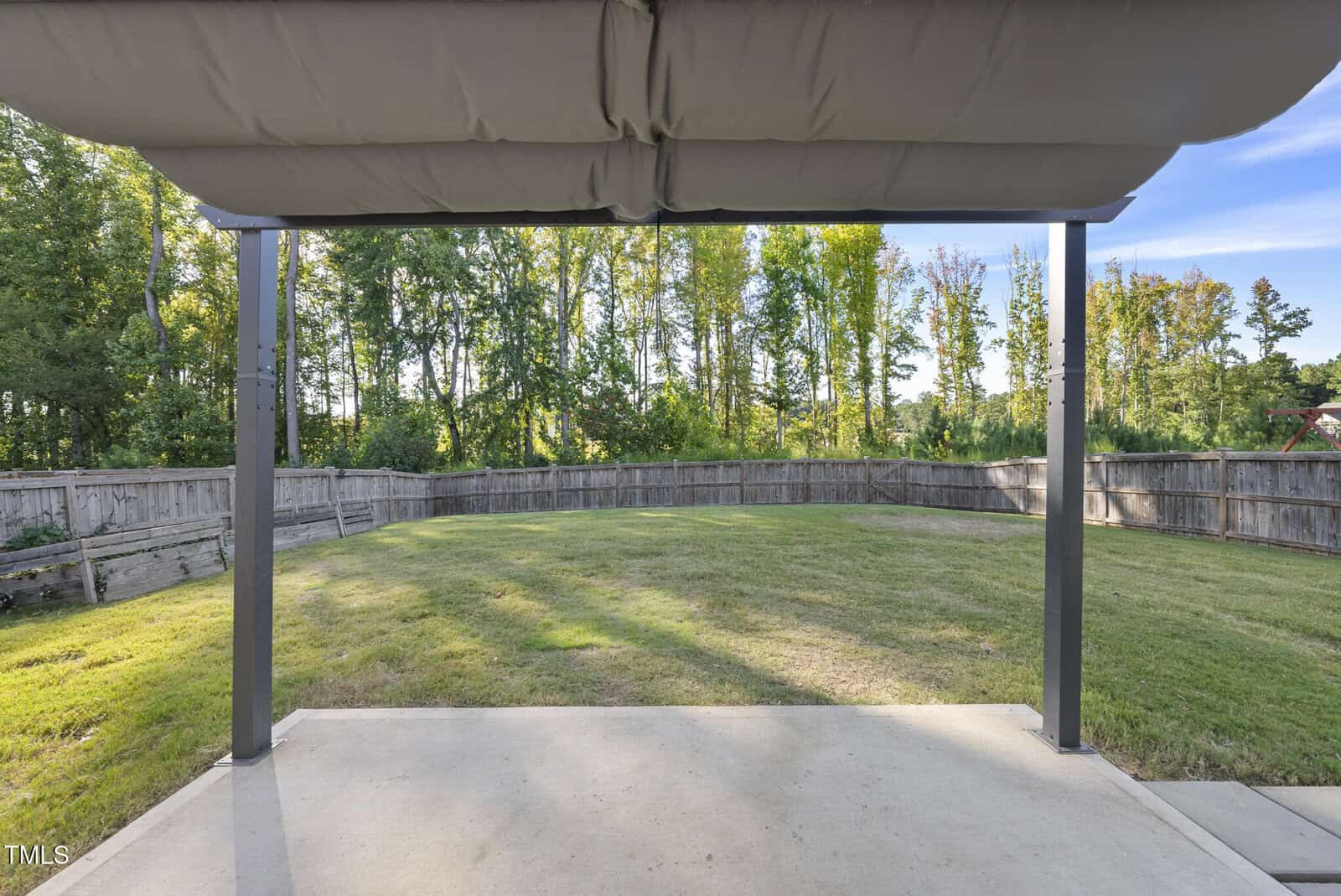 View from underneath a covered patio looking out onto a large, grassy backyard enclosed by a wooden fence, with tall trees lining the background under a clear blue sky.