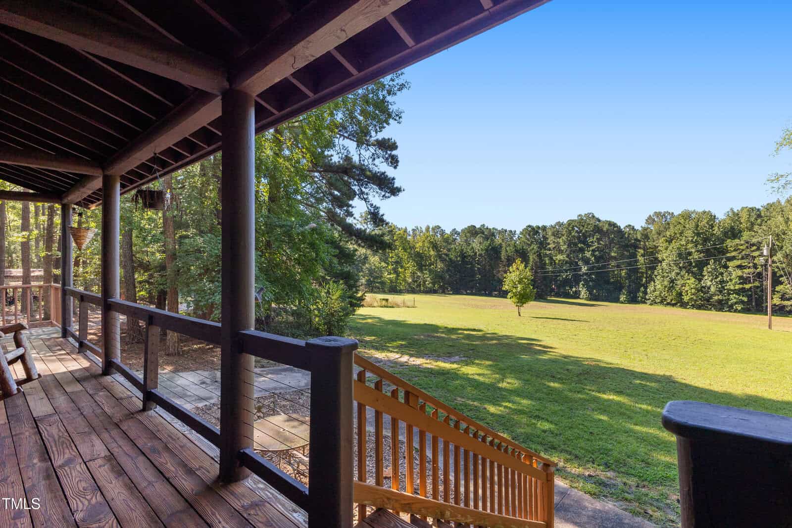A view from the wooden covered porch with railings at 104 Trails End Lane, Franklinton, looks out onto a grassy lawn, trees, and a clear blue sky. Wooden steps lead down from the porch to the yard.