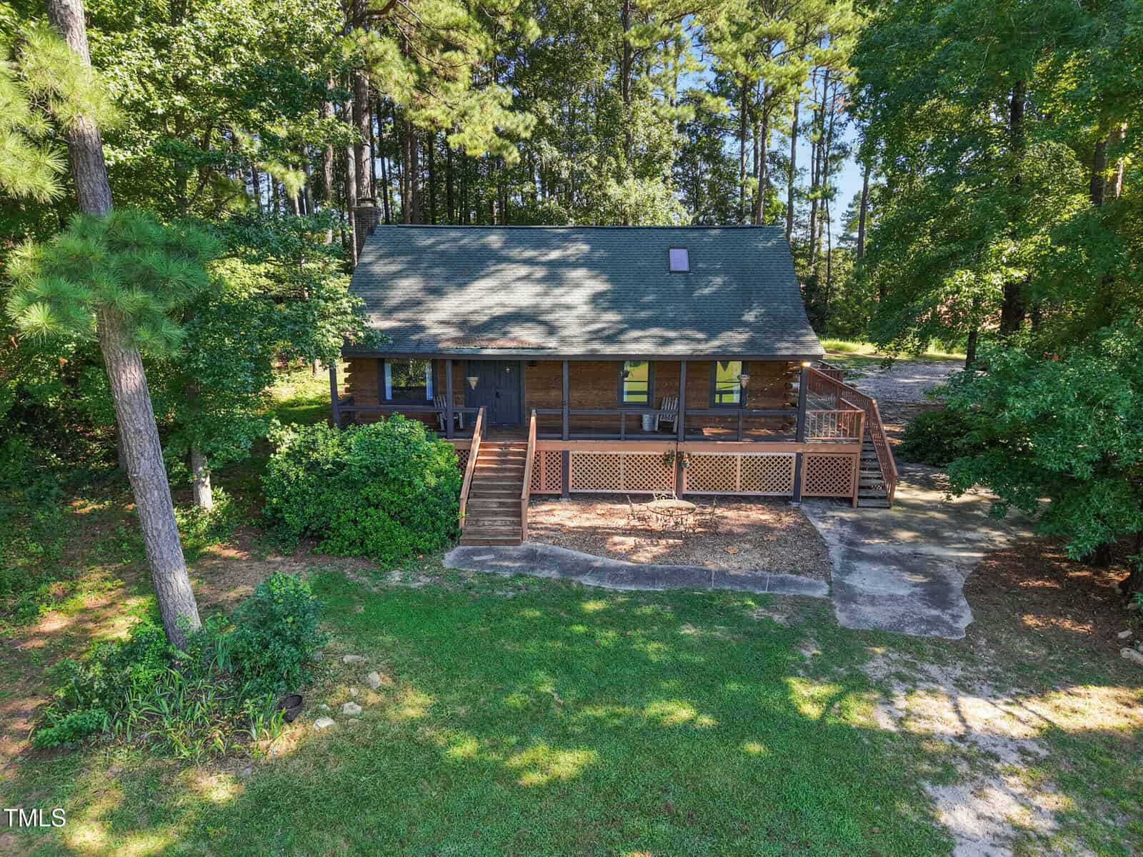 A log cabin with a covered front porch sits among tall trees. The house has a green roof, wooden railings, and a concrete walkway. Shadows from trees fall on the roof and yard. The lawn is partially grassy with some landscaping.