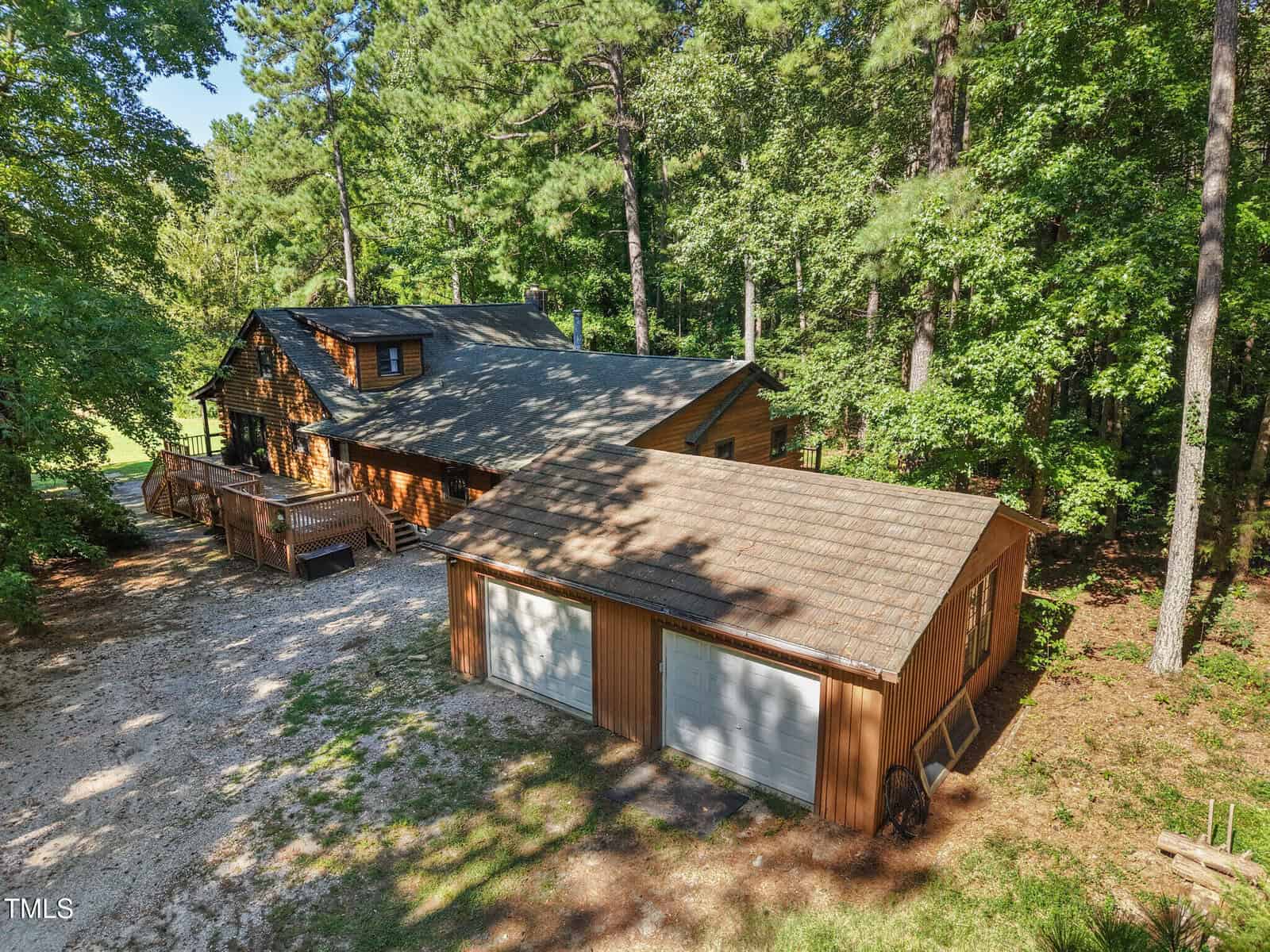 A wooden house with a large front deck is surrounded by trees. A separate two-car garage with matching wooden siding and a shingled roof sits in the foreground on a gravel driveway.