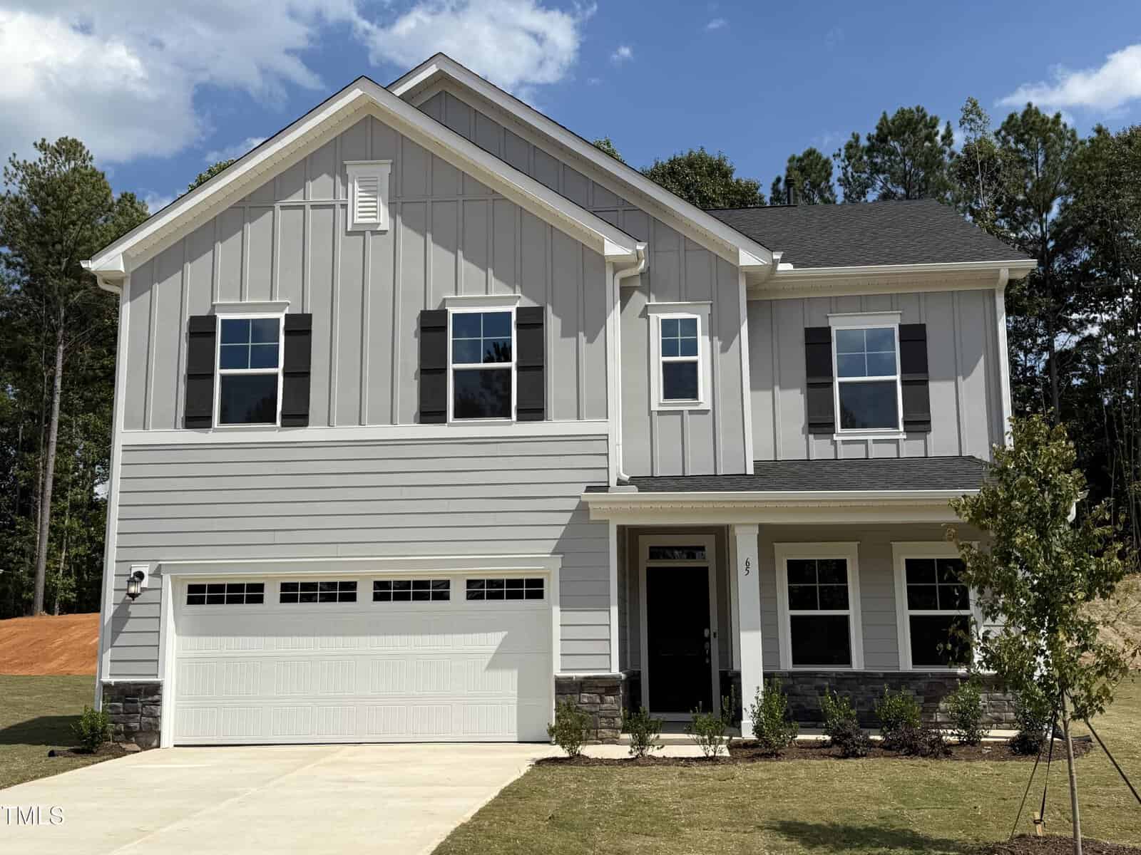 A modern two-story house with light gray siding, white trim, black shutters, and a double garage. The front yard has a small lawn, young trees, and shrubs, with a wooded area in the background.