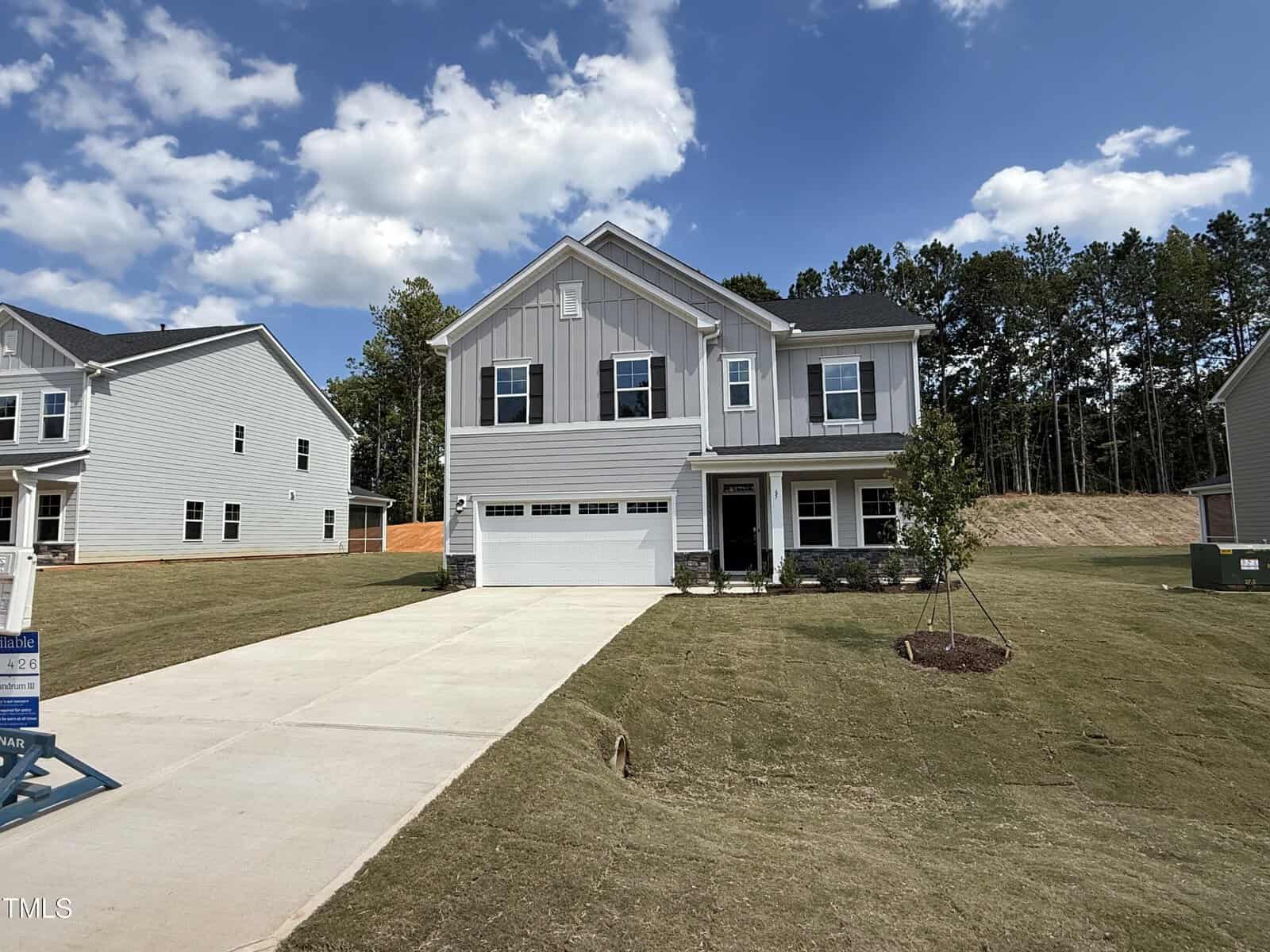 A modern two-story gray house with white trim and a double garage sits on a large, neatly manicured lawn. The driveway is wide, and the yard features a small tree. Tall trees line the background under a partly cloudy sky.