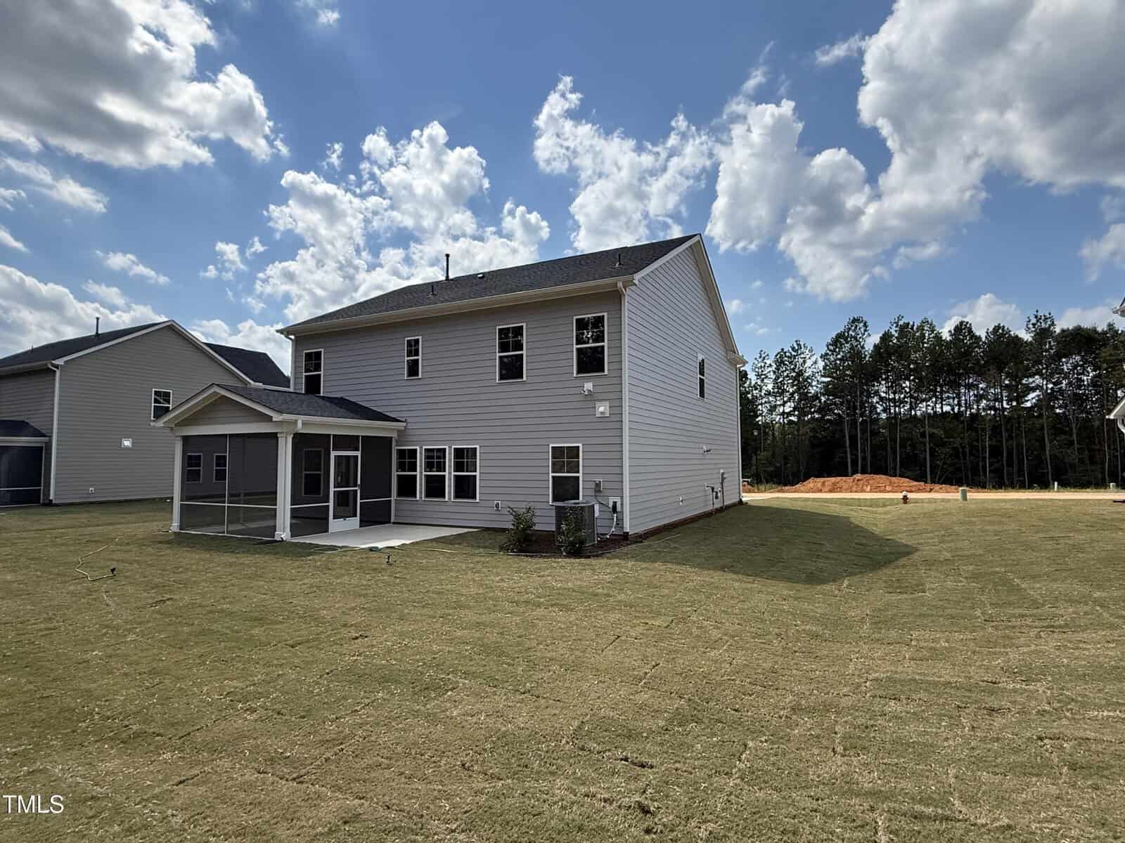 A two-story beige house with a screened patio and a large, freshly sodded backyard under a partly cloudy sky, surrounded by similar homes and a wooded area in the background.