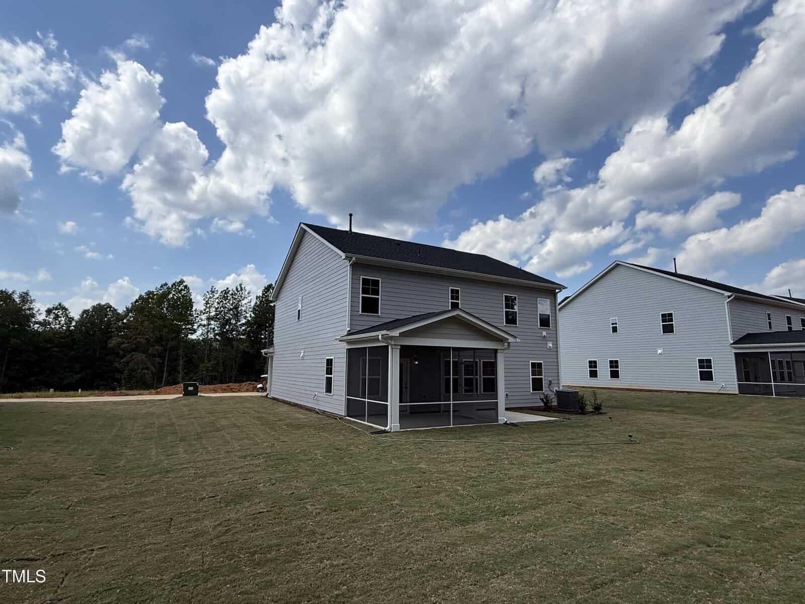A two-story, light gray house with a screened-in back porch sits on a large, grassy yard under a partly cloudy sky. Another similar house is visible in the background. Trees line the edge of the property.