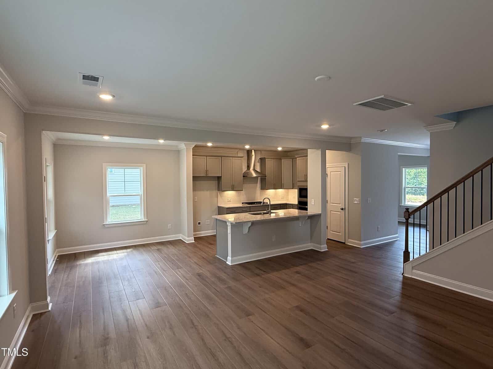 Open-concept kitchen and living area with wood flooring, neutral walls, recessed lighting, white cabinets, stainless steel appliances, and a staircase visible to the right. Large windows provide natural light.