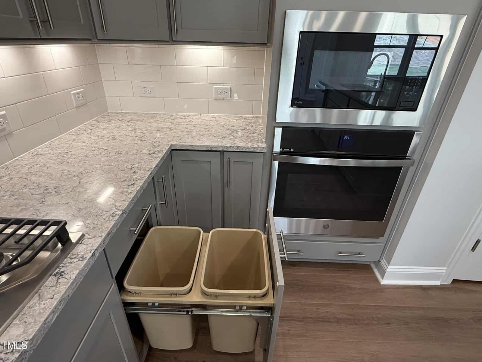A modern kitchen corner with gray cabinets, a built-in microwave and oven, marble countertop, gas stove, and a pull-out drawer containing two beige trash bins. The backsplash features white subway tiles with outlets.