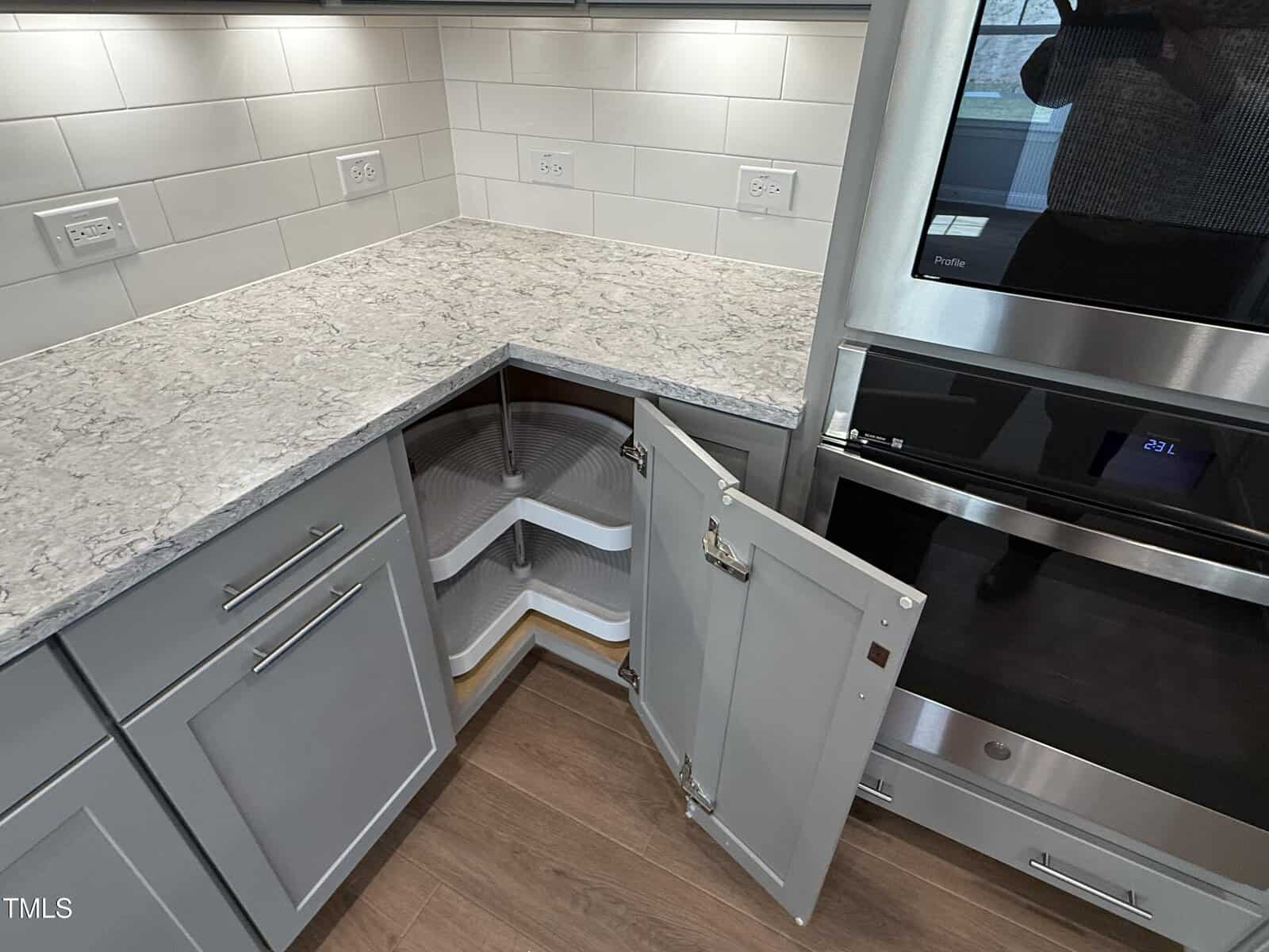 Corner kitchen cabinet with open doors revealing a white, two-tiered lazy Susan shelf. Gray cabinets, light granite countertop, white subway tile backsplash, and stainless steel appliance visible. Multiple wall outlets present.