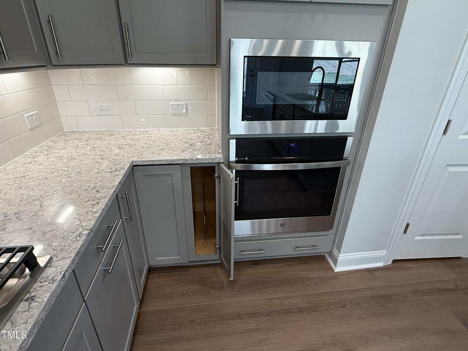 A modern kitchen features gray cabinets, marble-patterned countertops, stainless steel built-in microwave and oven, open lower cabinet door, tiled backsplash, and wood flooring. The countertop includes multiple electrical outlets.