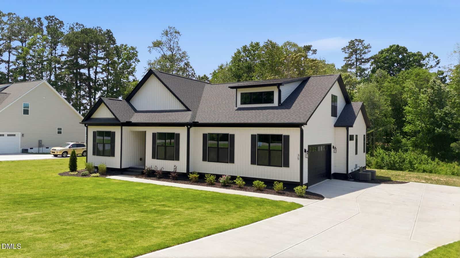 A modern, single-story house with white siding, black trim, multiple large windows, and a manicured lawn, driveway, and garage, surrounded by trees under a clear blue sky.