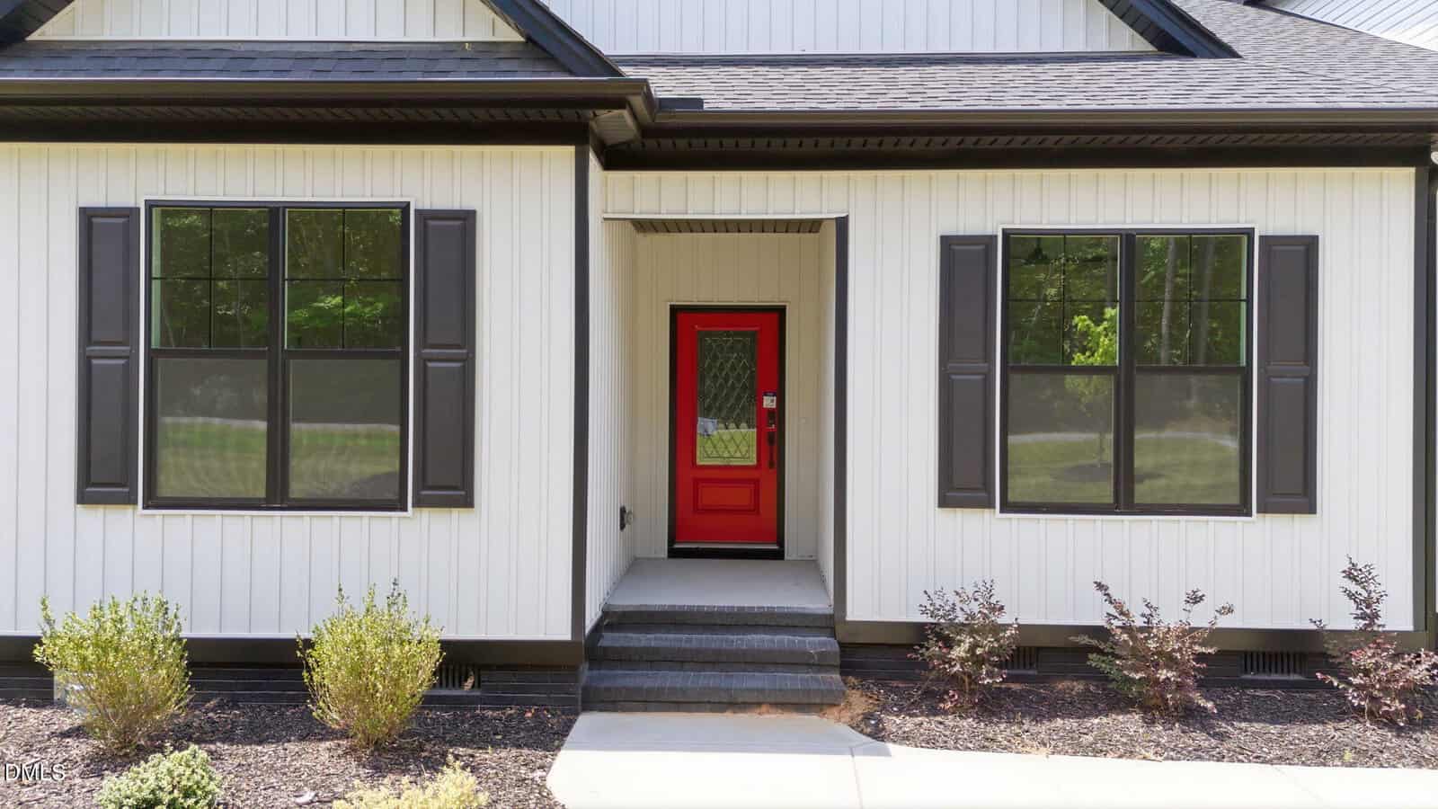 A modern house exterior with white vertical siding, black trim, and a bright red front door. Two large black-framed windows with shutters are on either side of the entry. Small shrubs and mulch line the walkway.