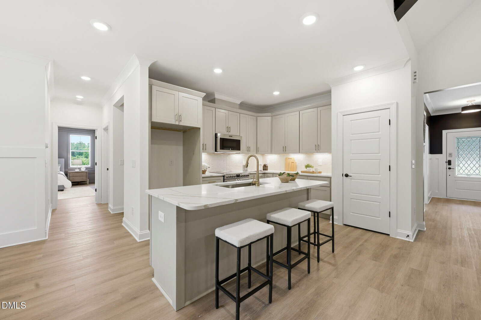 Modern kitchen with white cabinets, stainless steel appliances, marble island with sink, three cushioned stools, light wood flooring, and recessed lighting; open entryway and bedroom visible in the background.