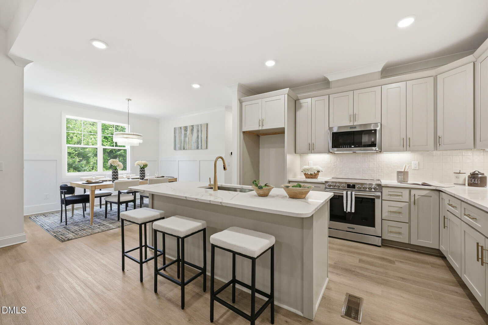 Modern kitchen with white cabinets, stainless steel appliances, a center island with three stools, light wood flooring, and an adjacent dining area with a large window and chandelier. The space is well-lit and uncluttered.