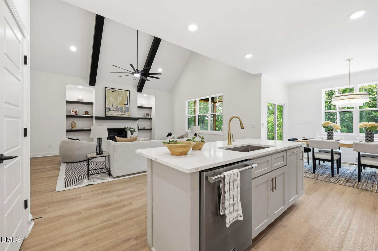 Open-concept modern living space with a kitchen island in the foreground, a living area with a couch and built-in shelves, and a dining area with windows overlooking greenery. White walls and light wood floors throughout.