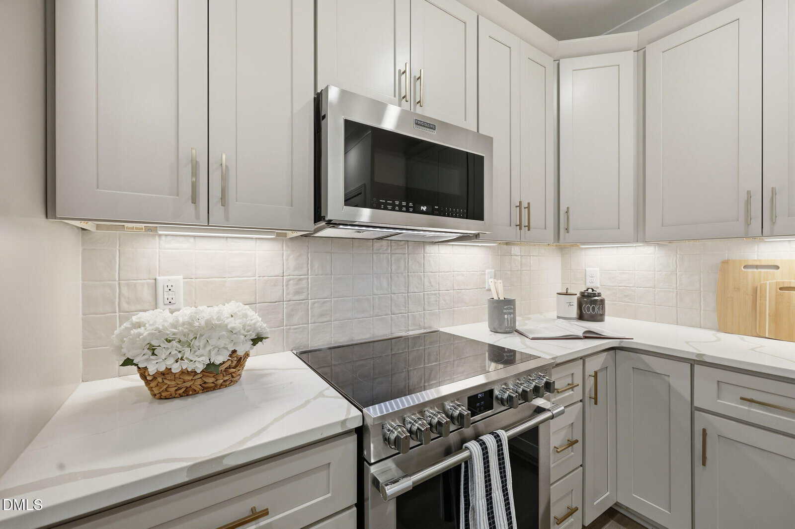 Modern kitchen with white cabinets, stainless steel oven and microwave, white tile backsplash, marble countertops, a basket of white flowers, and kitchen utensils neatly arranged on the counter.