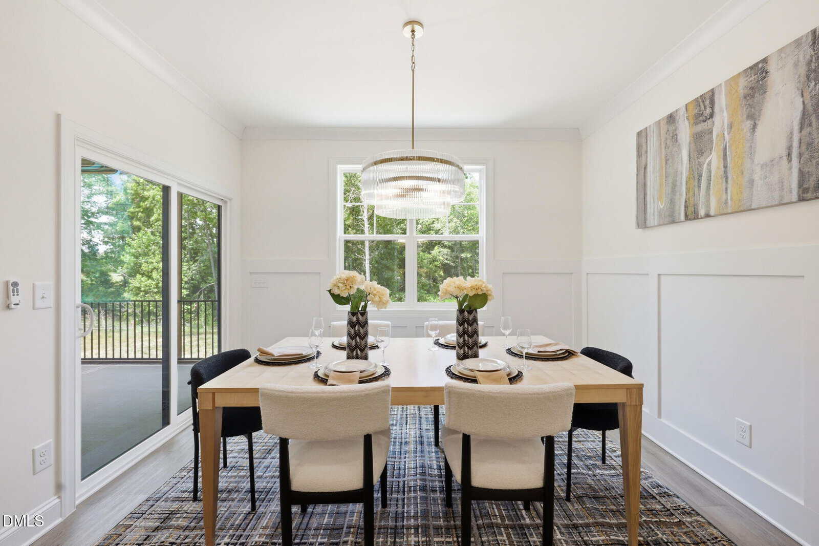A dining room with a light wood table set for six, beige upholstered chairs, black accent chairs at each end, a modern chandelier, large window, sliding door, and abstract art on the wall.