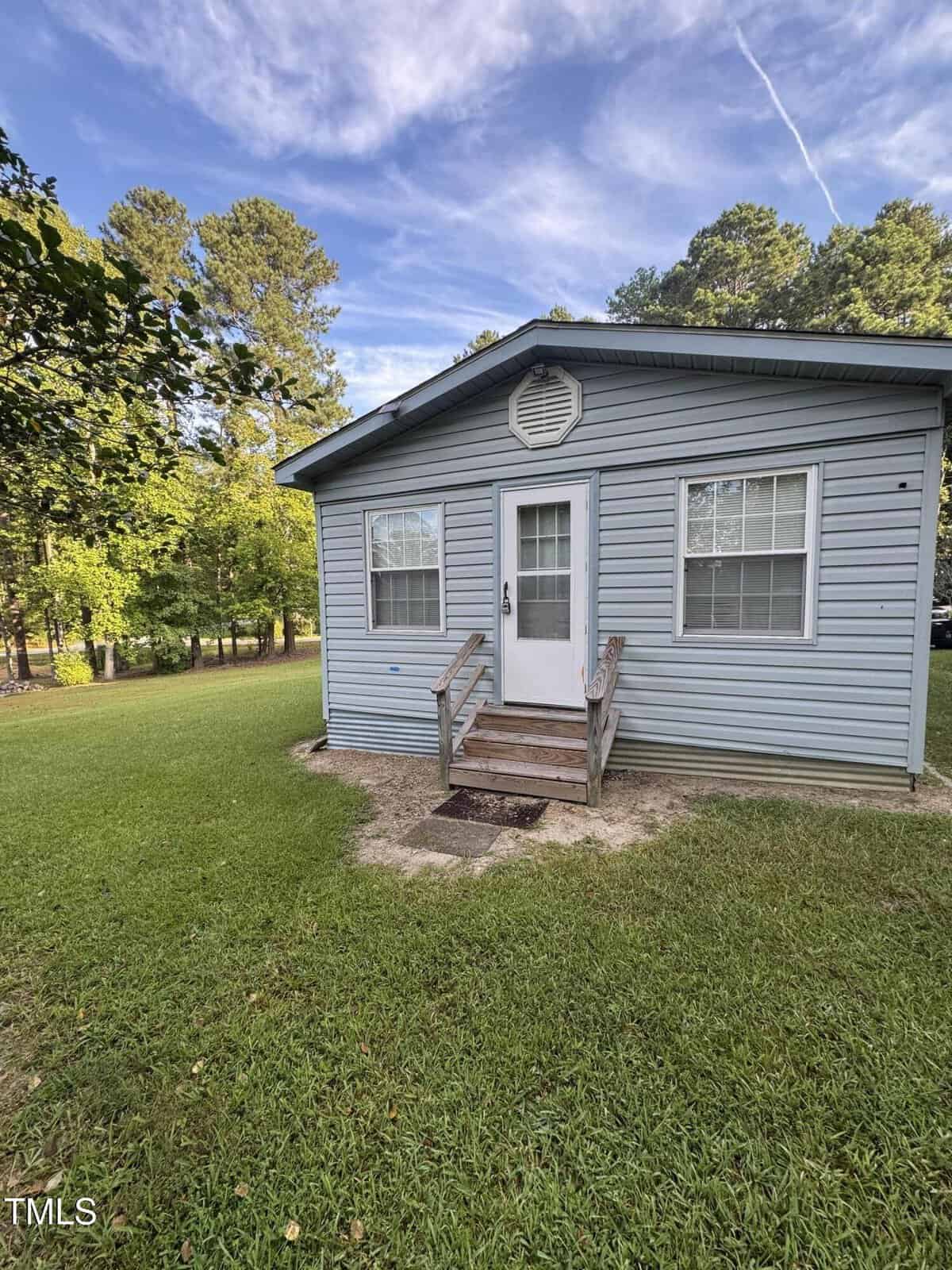 Charming small blue house with white trim and two windows, located at 108 Geronimo Drive in Louisburg. Set on a grassy yard, wooden steps lead to a welcoming white front door. Mature trees and a partly cloudy sky provide a picturesque backdrop.