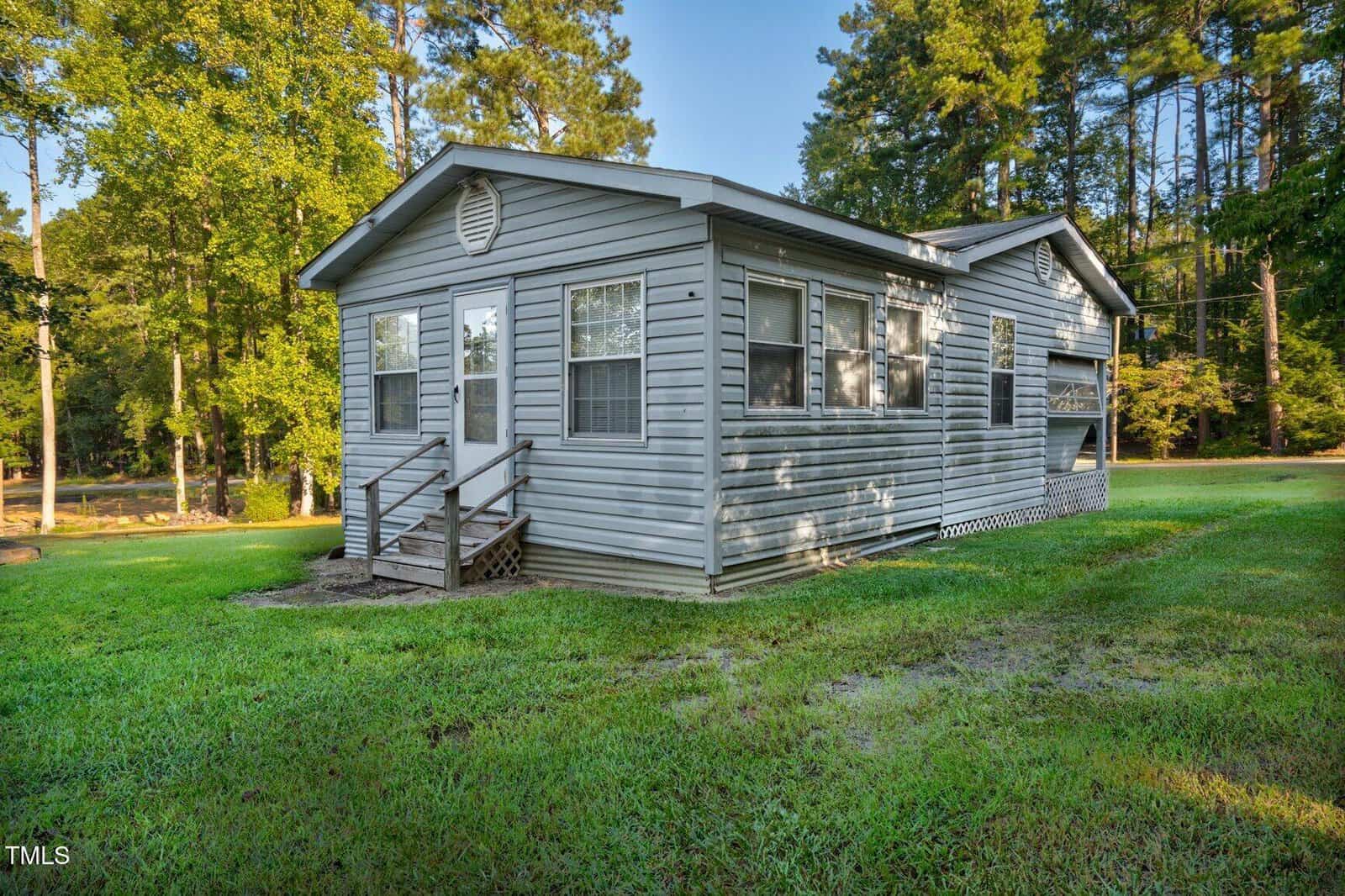 A small, single-story gray house with white trim sits on a grassy lawn, surrounded by tall trees. The house has multiple windows and a small wooden porch with steps leading to the front door.