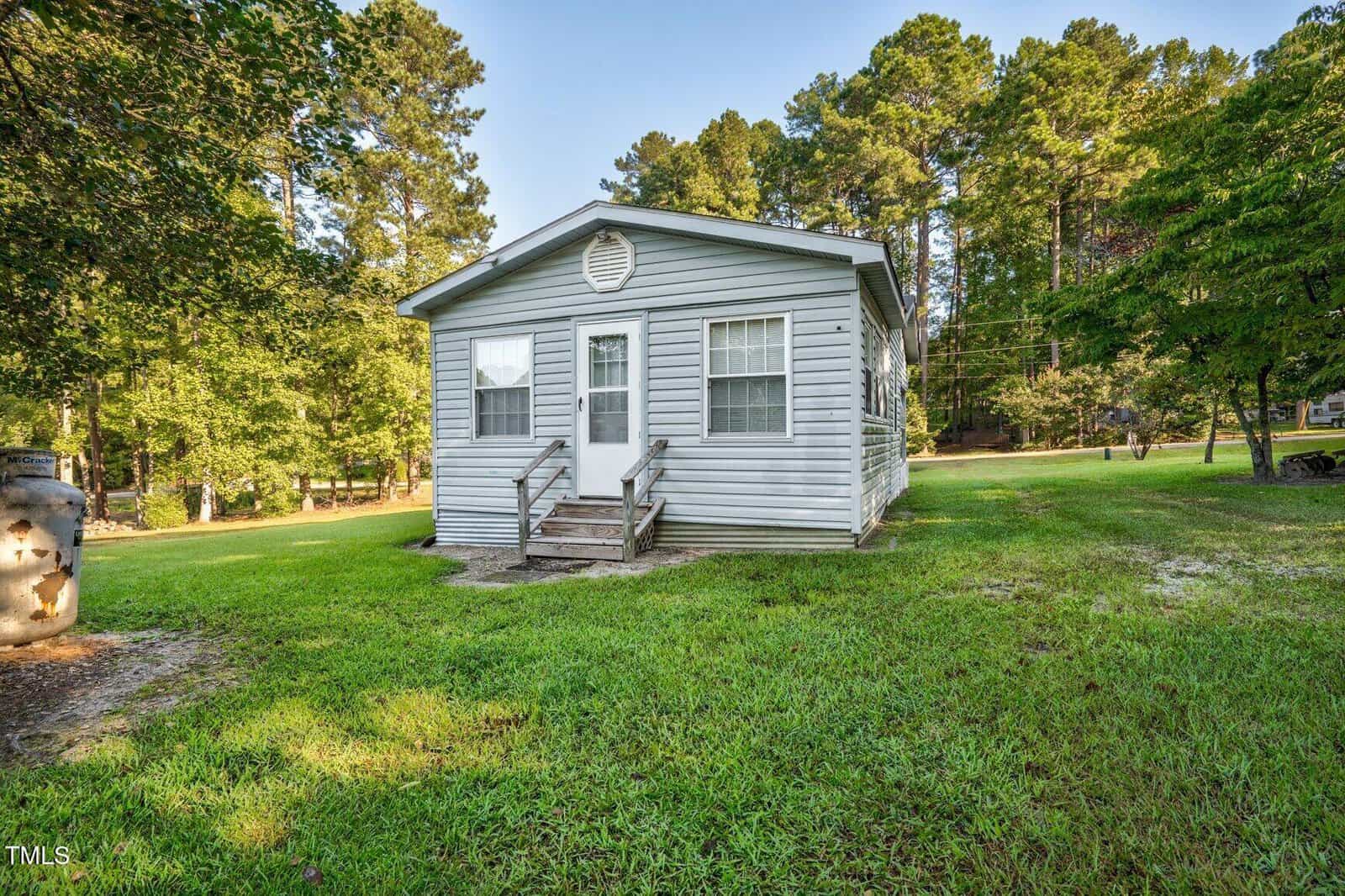 A small, single-story gray house with white trim and two front doors, sits on a grassy lawn surrounded by trees under a clear sky. Wooden steps lead up to each entrance.