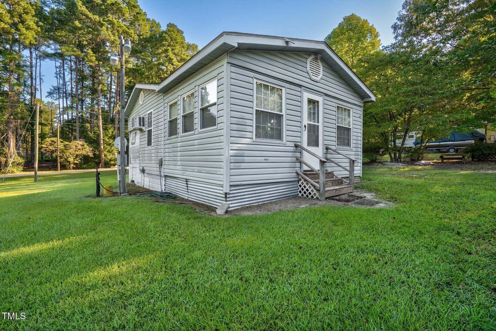 Small, single-story gray house with white trim, multiple windows, a small wooden front porch, and stairs. The house sits on a grassy lawn surrounded by trees. A parked car and utility poles are visible in the background.