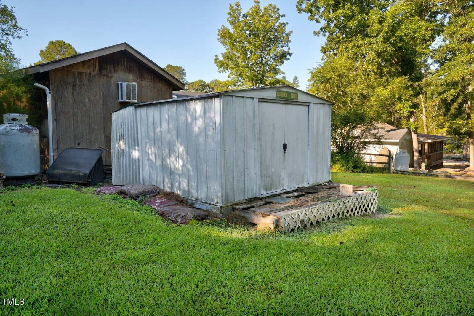 A metal storage shed with a closed door sits on a grassy lawn next to a wooden building. There are propane tanks, lattice skirting, and trees in the background under a clear sky.