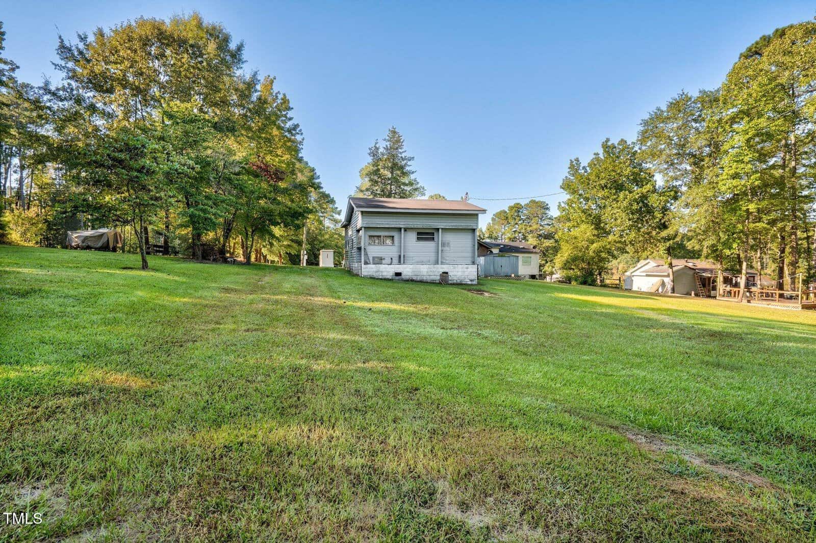 A small white house with a covered porch sits on a large, grassy lawn surrounded by trees. Outbuildings and more trees are visible in the background under a clear sky.