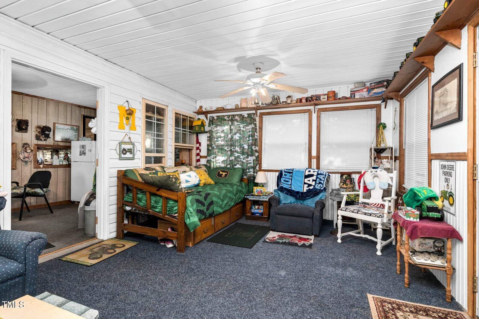 Cozy room with a green bed, blue armchair, and nature-themed decor. Windows let in light; shelves hold books and knickknacks. An open doorway leads to another room. White ceiling with a fan, dark blue carpet.
