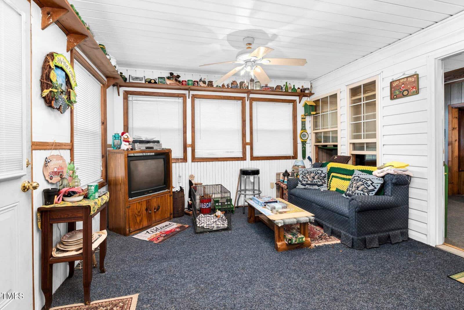 Cozy sunroom with blue carpet, a navy couch, wooden furniture, vintage TV, pet crates, ceiling fan, and knick-knacks on shelves and window ledges. Walls and ceiling are white, with decor giving a lived-in feel.