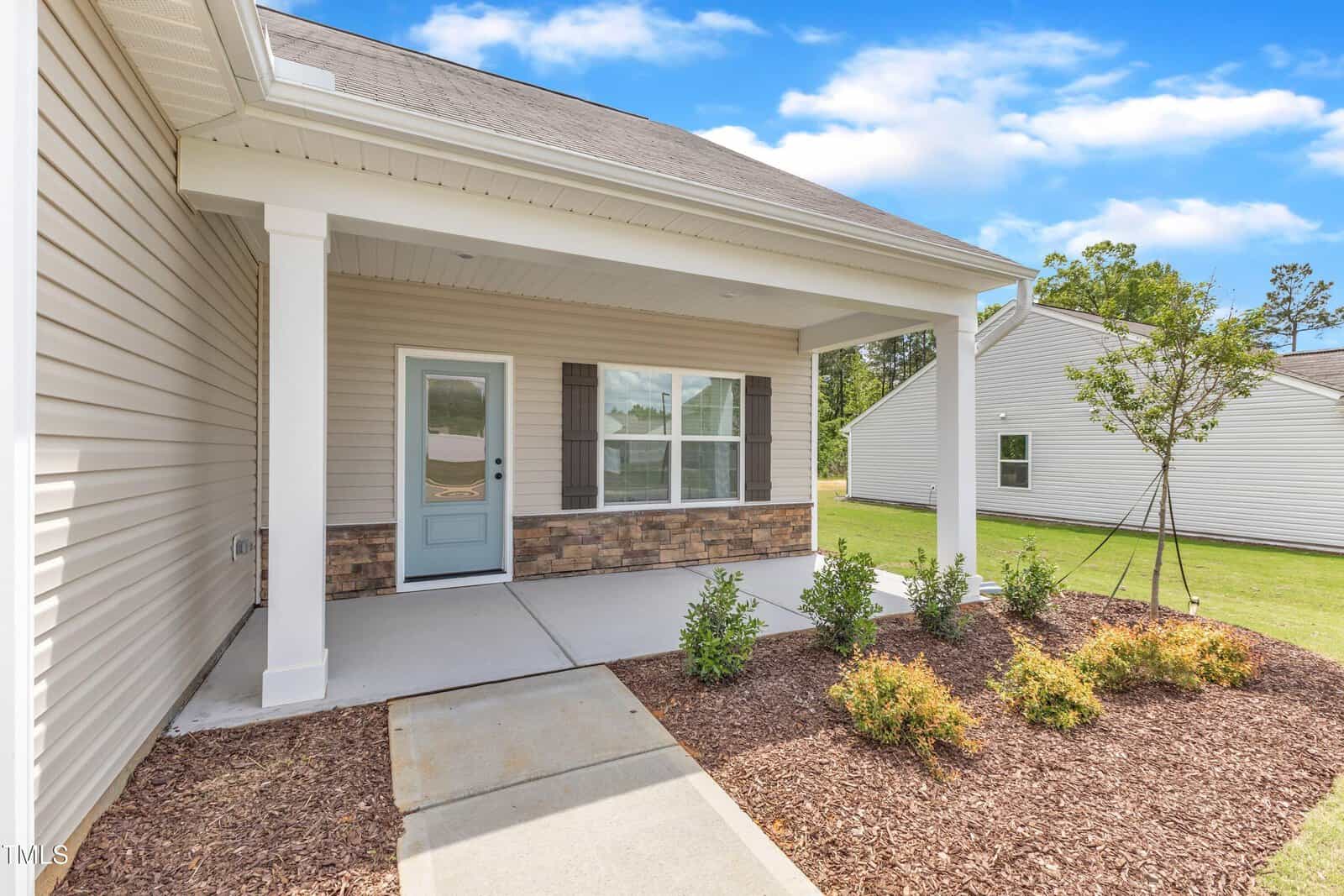 A covered front porch with white pillars, a light blue door, beige siding, and a landscaped area with small shrubs in front of a single-story house on a clear day.