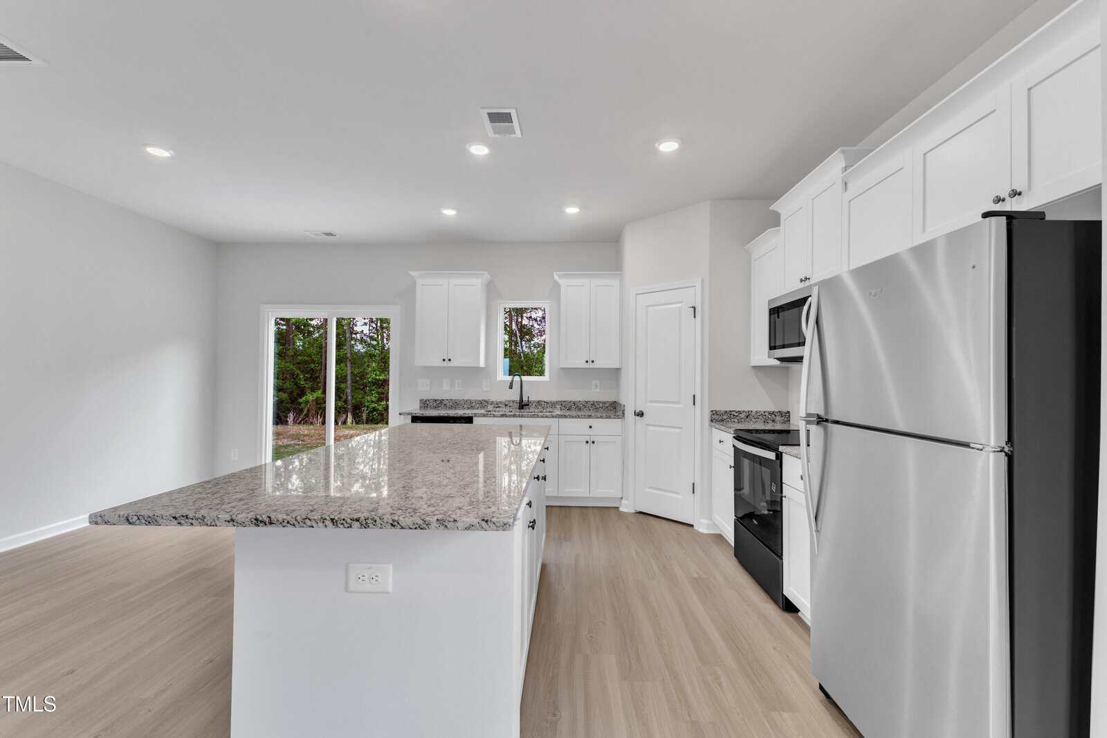 Modern kitchen with white cabinets, stainless steel appliances, granite countertops, a large island with outlets, and light wood flooring. Double doors open to an outdoor area. The room is brightly lit with recessed lighting.