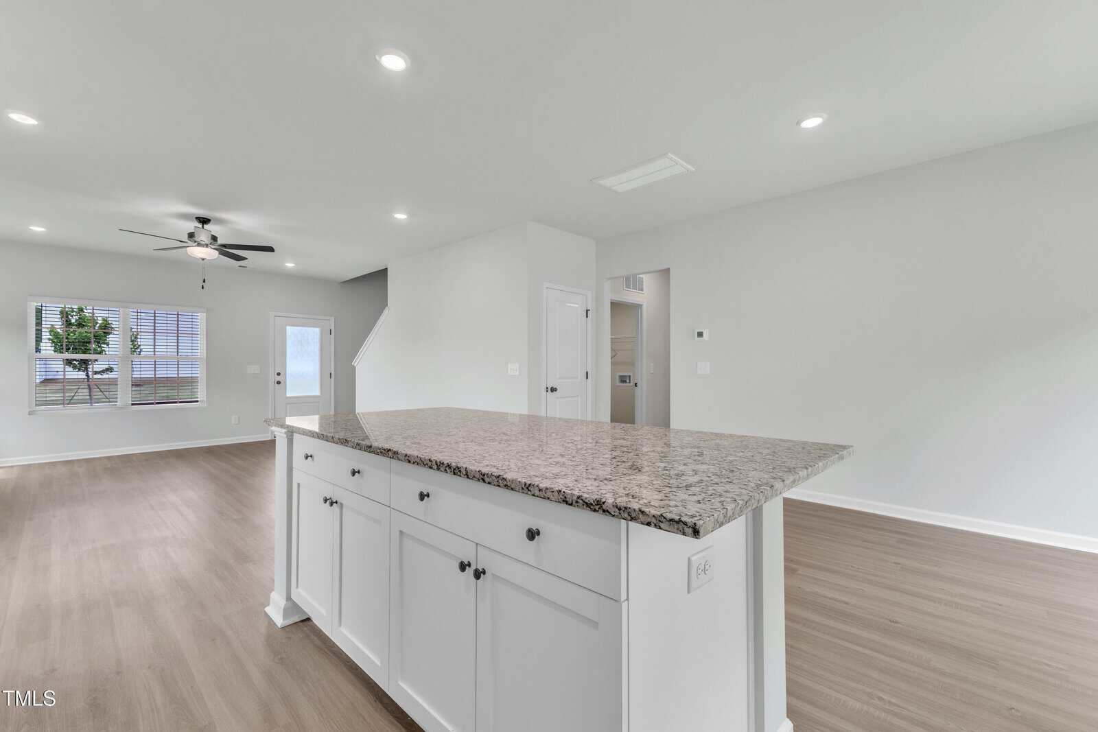 Spacious, modern open-concept living area with wood floors, white walls, a large granite kitchen island, ceiling fan, recessed lighting, and windows letting in natural light.