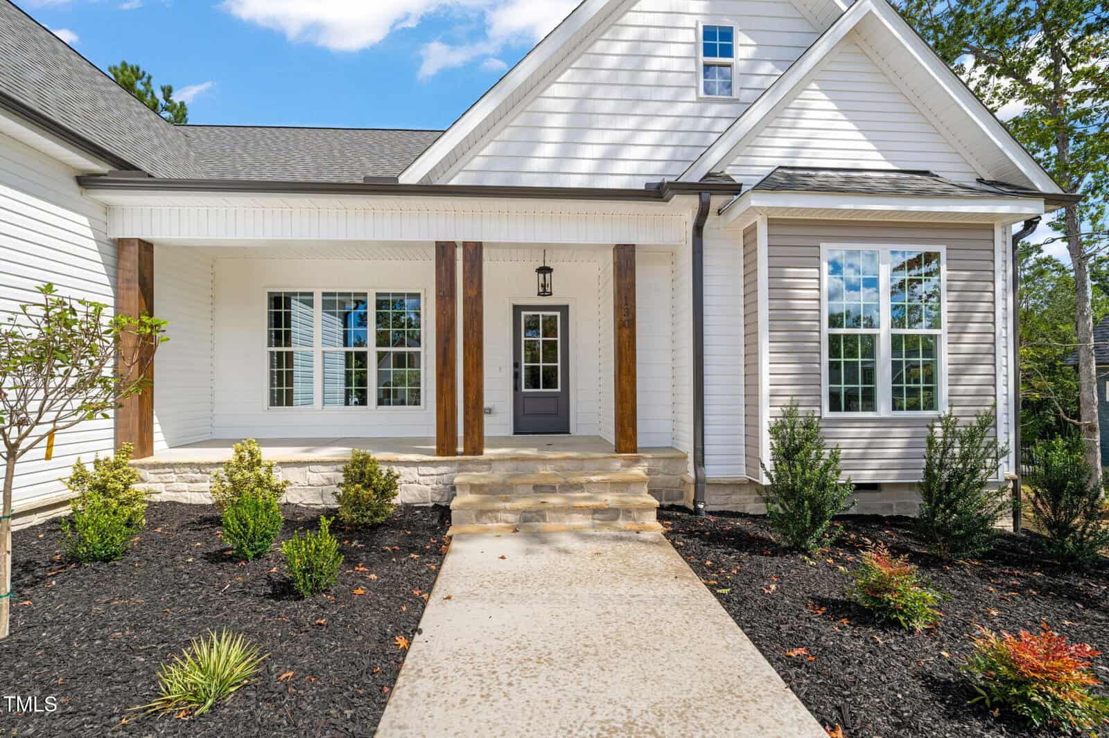 A modern single-story house with light-colored siding, a covered front porch supported by wooden columns, and landscaped shrubs bordered by concrete steps and a walkway.