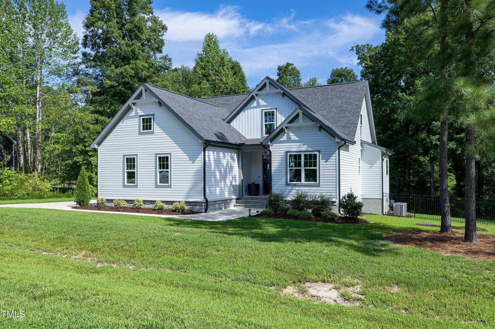 A modern white house with black trim sits on a large green lawn, surrounded by trees. The home features multiple gables, a covered front entry, and neatly landscaped bushes along the front.