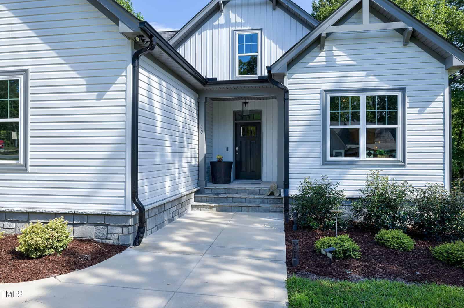 A modern white house with black trim, a covered entryway, gray front door, and landscaped shrubs along a concrete walkway in bright daylight.